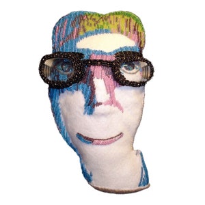 Può includere: Un ritratto di una persona con gli occhiali realizzato con perline. Il ritratto è fatto con perline colorate ed è cucito su uno sfondo di tessuto bianco.