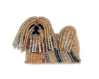May include: A beaded brooch shaped like a long-haired dog, crafted with gold, silver, and dark gray glass beads. The dog features a black bead nose and a textured, layered appearance created by the intricate beadwork. The text "the lone beader" is visible.