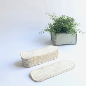 May include: A pile of beige, rectangular cloth pads with a textured surface and stitched edges. One pad is separate. A small, green plant in a concrete planter is in the background.