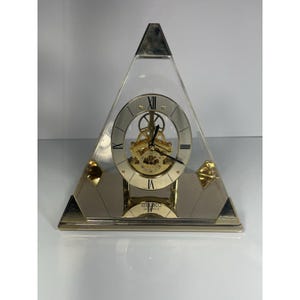 May include: A pyramid-shaped Seiko quartz clock with a clear front, revealing the inner workings. The clock face has Roman numerals and gold-colored accents. The base and top are silver-toned.