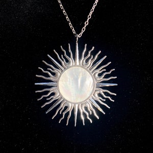 Puede incluir: Un collar de plata con un gran colgante de sol. El sol est&aacute; hecho de plata con un centro de n&aacute;car blanco.