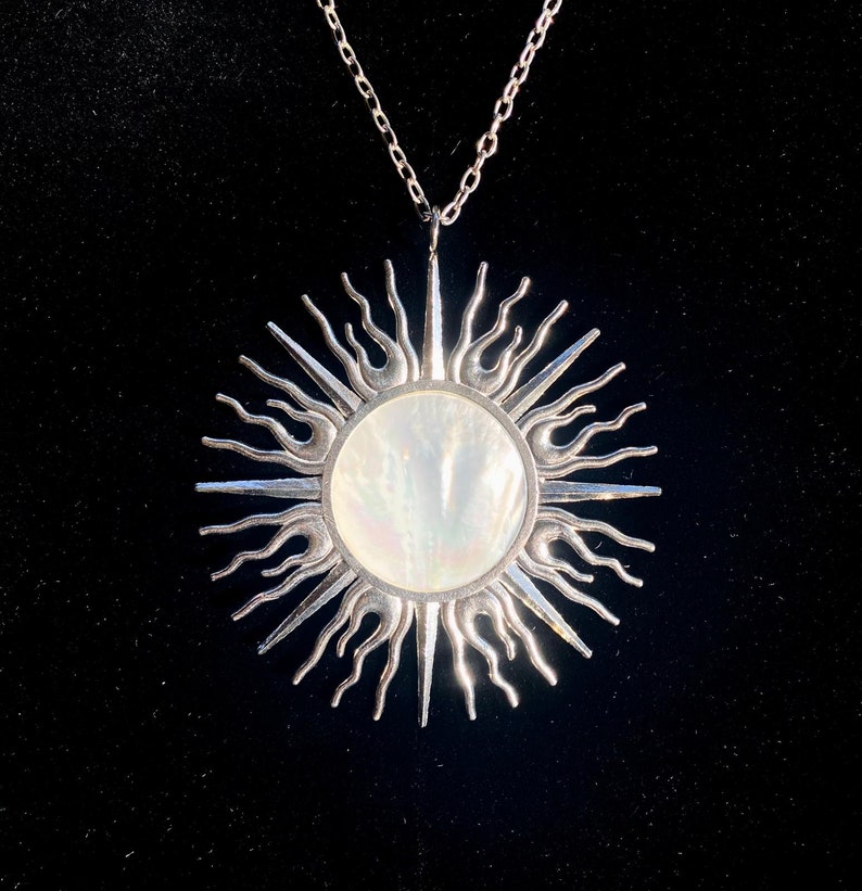 Puede incluir: Un collar de plata con un gran colgante de sol. El sol est&aacute; hecho de plata con un centro de n&aacute;car blanco.