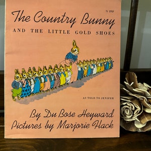 May include: A vintage children's book cover with a pink background. The title is "The Country Bunny and the Little Gold Shoes." The cover features an illustration of a line of bunnies wearing clothes and shoes.