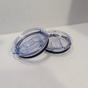 May include: Two clear plastic tumbler lids with black rubber seals. The lids have a sliding mechanism to open and close the drinking hole. The lids are oval-shaped and designed to fit a tumbler.