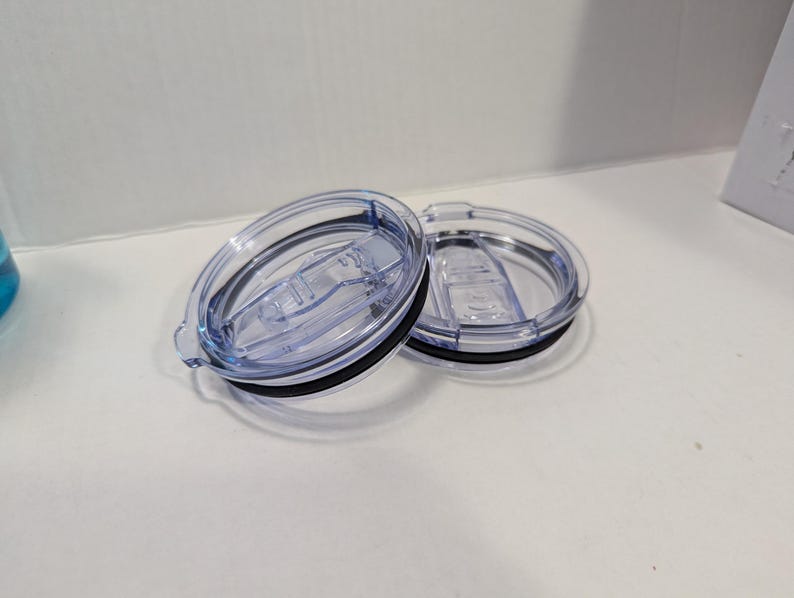 May include: Two clear plastic tumbler lids with black rubber seals. The lids have a sliding mechanism to open and close the drinking hole. The lids are oval-shaped and designed to fit a tumbler.