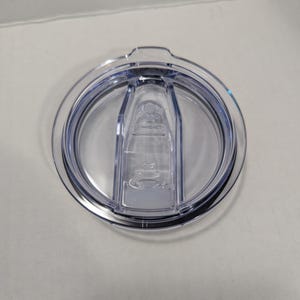 May include: Clear plastic tumbler lid with a sliding closure. The lid is circular with a raised center section and a dark rim. The sliding closure is transparent and has a small opening for drinking.