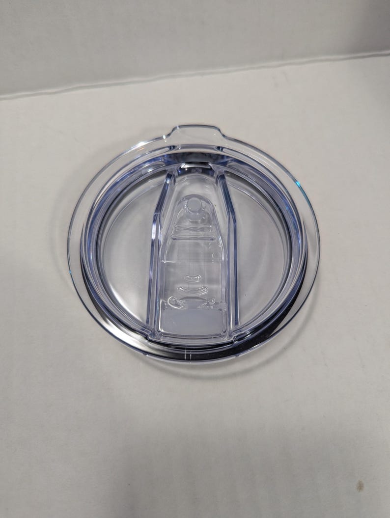 May include: Clear plastic tumbler lid with a sliding closure. The lid is circular with a raised center section and a dark rim. The sliding closure is transparent and has a small opening for drinking.