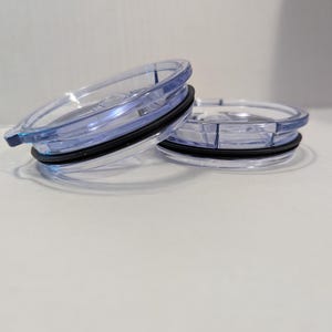 May include: Two clear plastic tumbler lids with a blue tint and black rubber seals. The lids are circular with a raised edge and a small tab for opening. One lid is slightly angled, revealing the design.