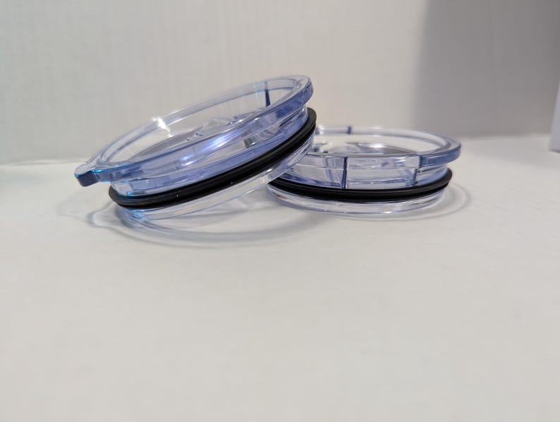 May include: Two clear plastic tumbler lids with a blue tint and black rubber seals. The lids are circular with a raised edge and a small tab for opening. One lid is slightly angled, revealing the design.