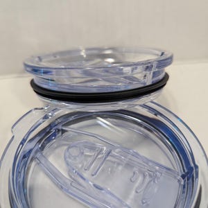 May include: Clear plastic tumbler lid with a black rubber seal and a sliding closure. The lid is designed to fit a travel mug or similar container. The lid is transparent, allowing the contents to be seen.