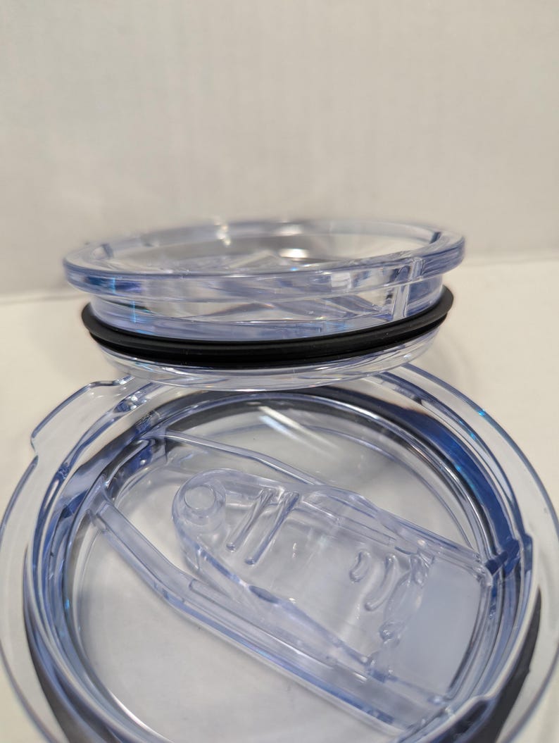 May include: Clear plastic tumbler lid with a black rubber seal and a sliding closure. The lid is designed to fit a travel mug or similar container. The lid is transparent, allowing the contents to be seen.