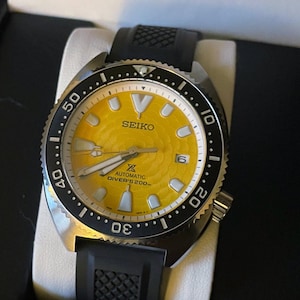 May include: A Seiko automatic diver's watch with a yellow dial and black bezel. The watch features a black rubber strap and a date window at the 3 o'clock position. The watch is presented in a black box.