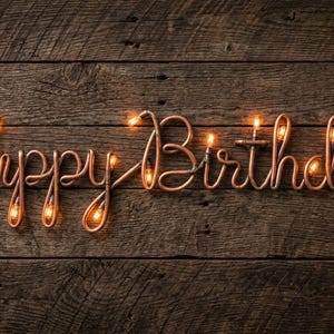 Peut inclure: Sculpture en fil de cuivre avec l'inscription "Happy Birthday!" éclairée par de petites lumières. Le texte aux tons chauds se détache sur un fond en bois rustique et vieilli, créant un visuel festif et accueillant.