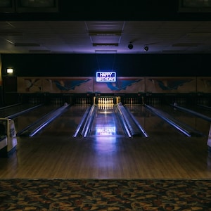 May include: A bowling alley interior featuring multiple wooden lanes, orange plastic seating, and a glowing blue neon sign that reads HAPPY BIRTHDAY suspended above the pins. The scene is dimly lit with reflective surfaces and vintage scoring consoles.