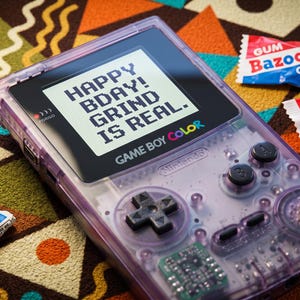 Può includere: Una console di gioco portatile Game Boy Color viola traslucido con uno schermo che mostra il testo HAPPY BDAY! GRIND IS REAL. Il dispositivo presenta pulsanti neri e un pad direzionale, accompagnato da vari involucri di caramelle tra cui Bazooka, Starburst e Jolly Rancher.