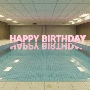 Puede incluir: Una piscina con las palabras "HAPPY BIRTHDAY" en letras rosas reflejadas en el agua. La piscina tiene azulejos azules y está rodeada por una habitación beige con paneles de madera y luces fluorescentes.