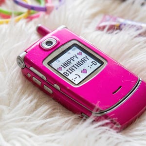 Könnte beinhalten: Ein leuchtend pinkes Klapphandy mit einem kleinen Display, das den Text HAPPY BIRTHDAY zusammen mit Herz- und Smiley-Emoticons anzeigt. Das Gerät verfügt über ein Kameraobjektiv, silberne Seitentasten und eine kleine Antenne an der Oberkante.