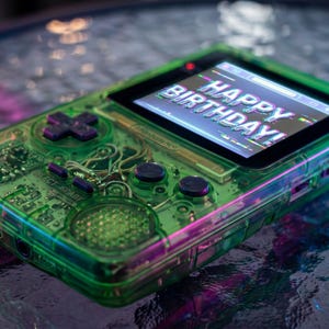 May include: A handheld video game console with a translucent neon green plastic shell, featuring a black directional pad, two black circular buttons, and a screen displaying the text HAPPY BIRTHDAY in a stylized, glitch-effect white font.