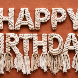 May include: A macramé wall hanging spelling out the words HAPPY BIRTHDAY in off-white knotted cord. Each letter is constructed from intricate woven patterns and features long, frayed fringe tassels hanging from the bottom of each character.