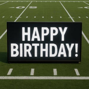May include: A black rectangular sign with the words "HAPPY BIRTHDAY!" in large white letters. The sign is on a green football field with white yard lines and markings.