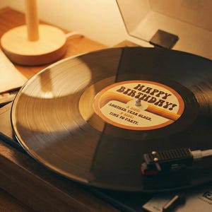 以下が含まれることがあります： 誕生日メッセージが書かれたレコードを再生するヴィンテージターンテーブル。レコードのラベルには「Happy Birthday! Another Year Older, Time to Party」と書かれています。ろうそくが灯ったカップケーキが近くにあり、レコードの山とランプが背景にあります。