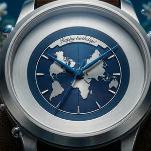 May include: A silver-toned wristwatch featuring a deep blue dial with a silver world map graphic. The watch face includes the text Happy birthday! in a script font, thin blue hands, and a brushed metal bezel with a crown on the right side.