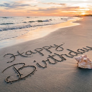Op de afbeelding: De woorden Happy Birthday staan in het zand op een strand bij zonsondergang. Een grote, getextureerde, lichtgekleurde schelp en een kleine, gladde, felblauwe steen liggen naast de tekst op het natte zand bij de kustlijn van de oceaan.