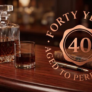 Può includere: Un decanter in vetro trasparente riempito con liquido ambrato, un bicchiere abbinato, un libro rilegato in pelle e una penna stilografica. Un emblema circolare color rame presenta il numero 40 e il testo FORTY YEARS AGED TO PERFECTION in disposizione circolare.