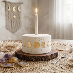 May include: A white cake with gold moon phase designs and a lit candle. The cake is on a wooden base, with crystals, dried plants, and a fork. The scene is set on a woven mat. The cake is approximately 10 cm in diameter.