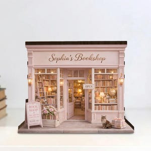 Puede incluir: Una maqueta rosa en miniatura de Sophia's Bookshop con detalles interiores y exteriores. La tienda tiene un letrero que dice "Open" y un pequeño gato afuera. Un letrero dice "Book Shop Come In".