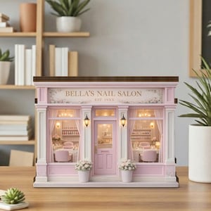 May include: A miniature pink nail salon model with the text "Bella's Nail Salon" above the door. The model has white columns, a brown roof, and small white flower pots. The interior is visible through the windows.