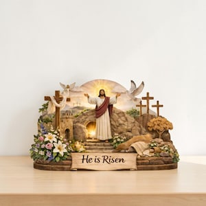 May include: A religious figurine showing Jesus with outstretched arms, rising from a tomb. The scene includes crosses, doves, and the text "He is Risen" on a banner. The figurine is surrounded by flowers and a glowing light.