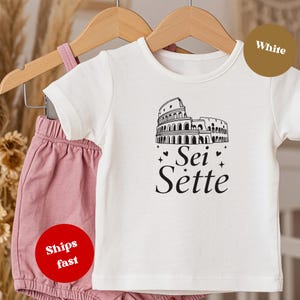 May include: White baby t-shirt featuring a black illustration of the Colosseum and the words "Sei Sette". The t-shirt is displayed with pink shorts on a wooden hanger.