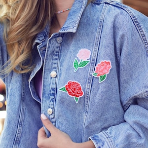 Könnte beinhalten: Hellblaue Jeansjacke mit drei Blumenaufnähern in Rosa und Rot. Die Jacke hat ein klassisches Knopfdesign und wird über einem lila Kleid getragen. Der Hintergrund zeigt eine verschwommene Szene eines Vergnügungsparks.