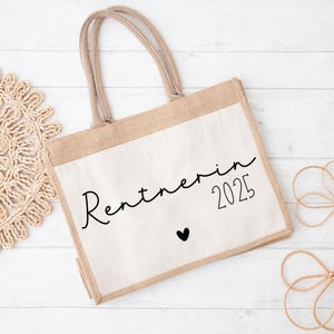 Könnte beinhalten: Beige Tragetasche mit weißer Frontplatte. Der Text "Rentnerin 2025" ist in schwarzer Schrift geschrieben, darunter ein kleines schwarzes Herz. Die Tasche hat zwei beige Griffe.