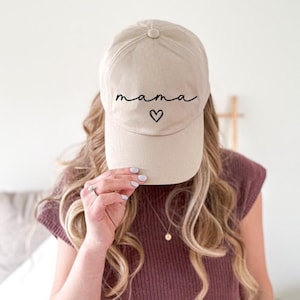 Könnte beinhalten: Beige Baseballkappe mit dem Wort "Mama" in schwarzer Schrift und einem Herzsymbol. Die Kappe hat einen gebogenen Schirm und einen Knopf oben. Der Hut ist neutralfarben und für den täglichen Gebrauch geeignet.