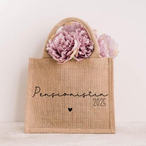 Könnte beinhalten: Eine beigefarbene Jutetasche mit Henkel, gefüllt mit rosa Blumen. Auf der Tasche steht das Wort "Pensionistin" und das Jahr "2025", sowie ein kleines Herzsymbol.