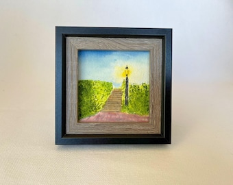 Könnte beinhalten: Ein gerahmtes Aquarell zeigt einen Weg zu einer Straßenlaterne, flankiert von grünen Hecken. Der Himmel ist blau mit einem Hauch von Gelb. Der Rahmen hat einen grauen Innenrand und einen schwarzen Außenrahmen.