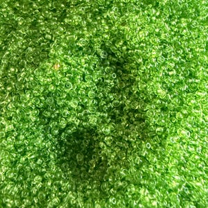 May include: A close-up view of a pile of small, translucent, lime green seed beads. The beads are densely packed, creating a textured surface. The color is vibrant and consistent throughout the image, suggesting a craft supply.