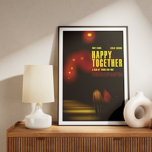 May include: A framed film poster for "Happy Together" with a black frame, featuring yellow text on a dark background. A white lamp and decorative vases sit on a wooden cabinet.