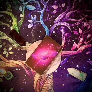 Pode incluir: Um baralho de cartas de tarot com um design rosa e roxo, repousando sobre uma superf&iacute;cie circular branca. Cristais e um padr&atilde;o de &aacute;rvore colorido em um fundo escuro cercam as cartas. Tema espiritual ou metaf&iacute;sico.
