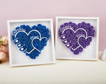 Può includere: Due composizioni floreali a forma di cuore incorniciate. Una presenta rose blu, l'altra viola. Ognuna ha una sovrapposizione a forma di cuore bianco con le parole "Nonna" e "Mamma", più nomi. Le cornici sono bianche.