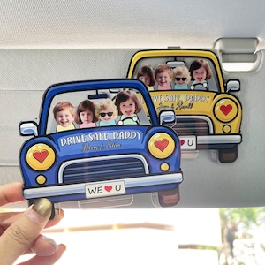 May include: Two car-shaped sun visors, one blue and one yellow, each featuring a photo of children. The blue car has the text "DRIVE SAFE DADDY" and "WE U". The yellow car also has the text "DRIVE SAFE DADDY". Both have heart accents.