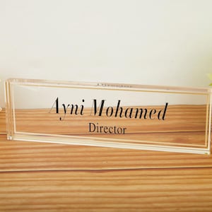 May include: A clear acrylic desk nameplate with gold trim, displaying the name "Ayni Mohamed" and the title "Director" in elegant black script. The nameplate rests on a wooden surface, with floral accents in the background.