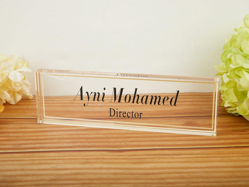 May include: A clear acrylic desk nameplate with gold trim, displaying the name "Ayni Mohamed" and the title "Director" in elegant black script. The nameplate rests on a wooden surface, with floral accents in the background.