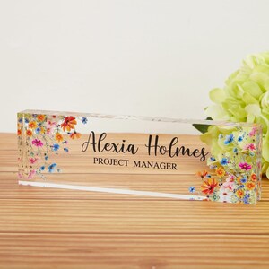 May include: A clear acrylic desk nameplate with floral accents. The nameplate features the name "Alexia Holmes" and the title "PROJECT MANAGER" in black text. Colorful flowers in shades of red, blue, and yellow decorate the edges.