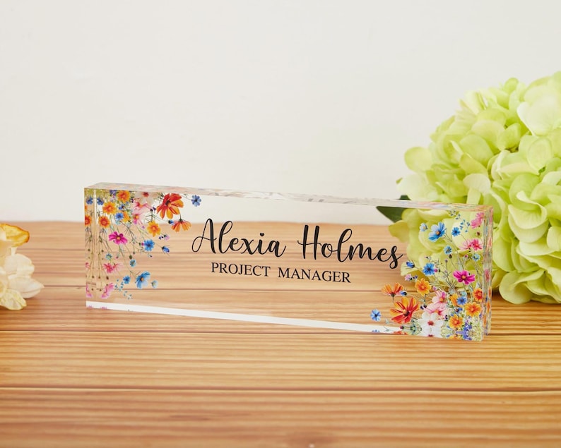May include: A clear acrylic desk nameplate with floral accents. The nameplate features the name "Alexia Holmes" and the title "PROJECT MANAGER" in black text. Colorful flowers in shades of red, blue, and yellow decorate the edges.