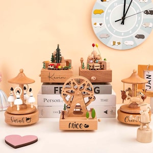Könnte beinhalten: Eine Sammlung von Holzspieluhren, darunter ein Riesenrad, ein Karussell und ein Zug-Set, mit eingravierten Namen wie "Sarah", "Kevin" und "Alice". Im Hintergrund hängt eine Uhr mit Meerschweinchen-Illustrationen.