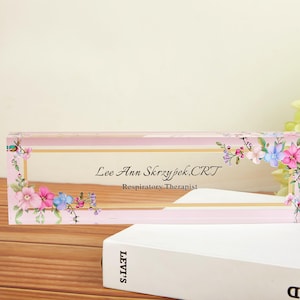 May include: A rectangular, clear acrylic desk nameplate with a pink border and floral accents. The nameplate features the name "Lee Ann Skrzypek, CRT" and the title "Respiratory Therapist". The design includes pink, blue, and purple flowers.