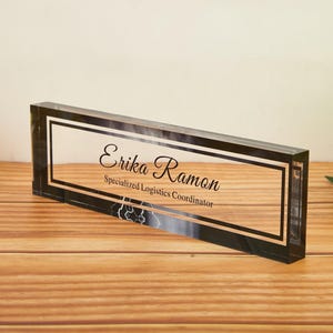 May include: A rectangular clear acrylic desk nameplate with a black border. The nameplate features the name "Erika Ramon" and the title "Specialized Logistics Coordinator" in black script.